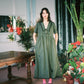 Woman in a green dress standing in a room with plants and a table.