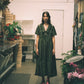 Woman in a green dress standing in a room with plants and furniture.