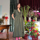 Woman standing in a mid length olive green dress with her hands in her pockets in a room with plants and flowers.