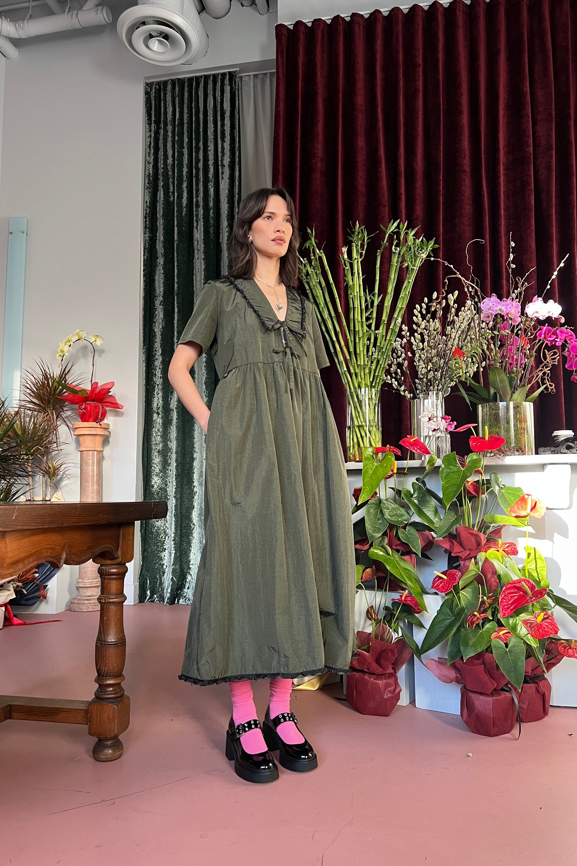 Woman standing in a mid length olive green dress with her hands in her pockets in a room with plants and flowers.