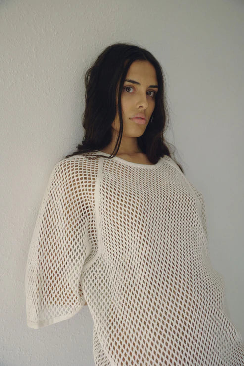 Close-up of Bohème Goods Fisherman Tee in cream showing open net cotton knit texture and relaxed fit, model leaning against a white wall