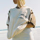 Bohème Goods Tourist Tee in cream worn on a beach with cream shorts, showing Club Bohème palm tree graphic and brown stripe sleeve detail with ocean in the background