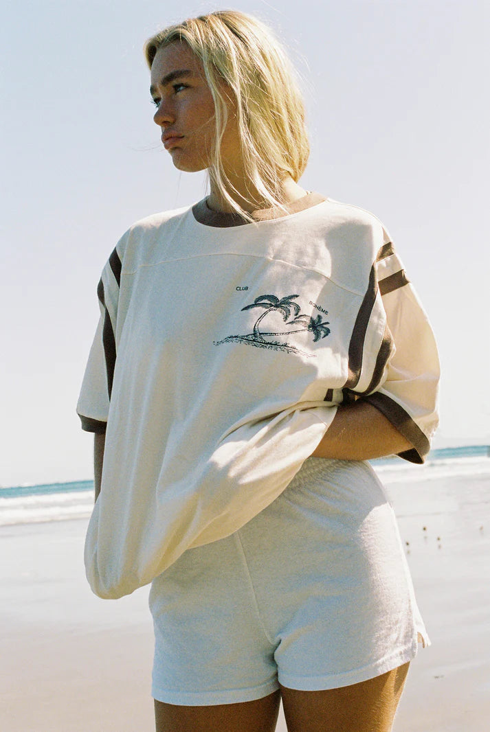Bohème Goods Tourist Tee in cream worn on a beach with cream shorts, showing Club Bohème palm tree graphic and brown stripe sleeve detail with ocean in the background