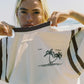 Close-up of Bohème Goods Tourist Tee in cream showing Club Bohème palm tree island graphic and brown contrast ribbed neckline, held open at beach with bright natural light