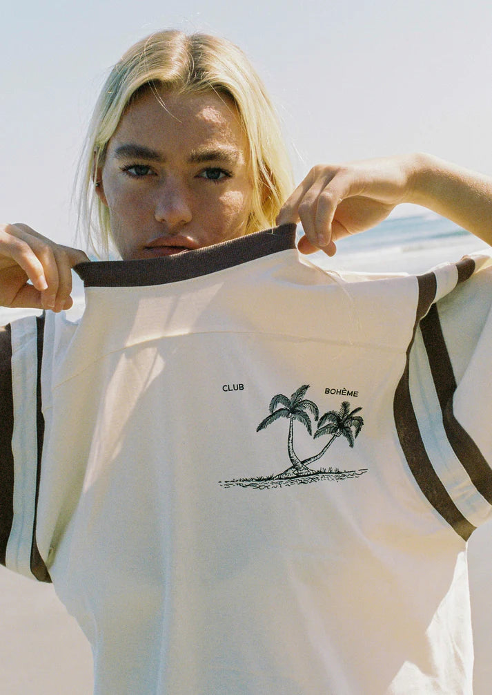 Close-up of Bohème Goods Tourist Tee in cream showing Club Bohème palm tree island graphic and brown contrast ribbed neckline, held open at beach with bright natural light