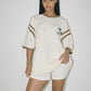 Bohème Goods Tourist Tee in cream styled with matching Track Short and tan cowboy boots, tee knotted at hip showing Club Bohème chest graphic and brown stripe sleeve detail