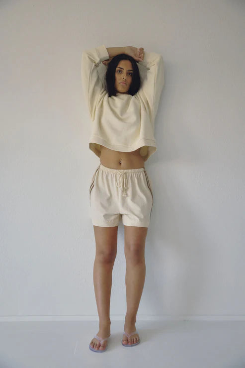 Bohème Goods Track Short in cream with brown side stripes worn with a cropped cream sweatshirt, arms raised, showing relaxed fit and drawstring waist against a white studio background