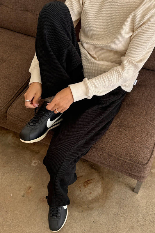 Le Bon Shoppe Waffle Lo Pants in true black worn seated on a brown sofa, styled with the cream Waffle Fundamental Tee and black sneakers, showing heavy waffle cotton texture