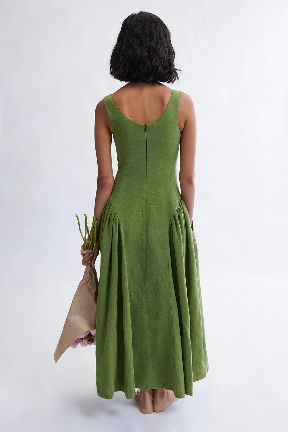Woman wearing a meadow green linen midi dress with scoop neckline and voluminous panier skirt, holding pink peonies — Vivienne Dress by Eliza Faulkner at Ånde