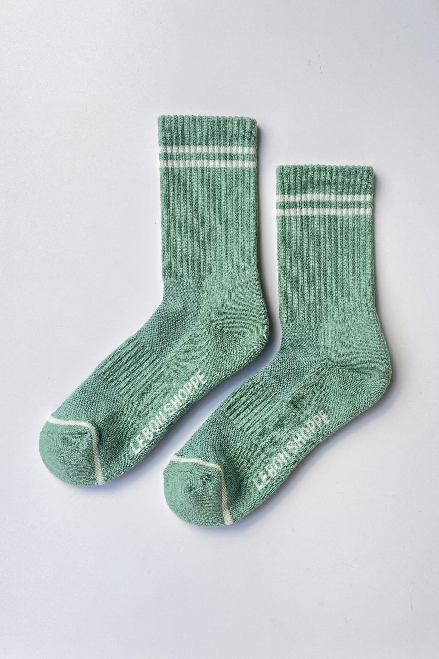Pair of Le Bon Shoppe Boyfriend Socks in meadow laid flat showing sole with mesh panel, Le Bon Shoppe branding, contrast toe detail and white double stripe on green ribbed cotton