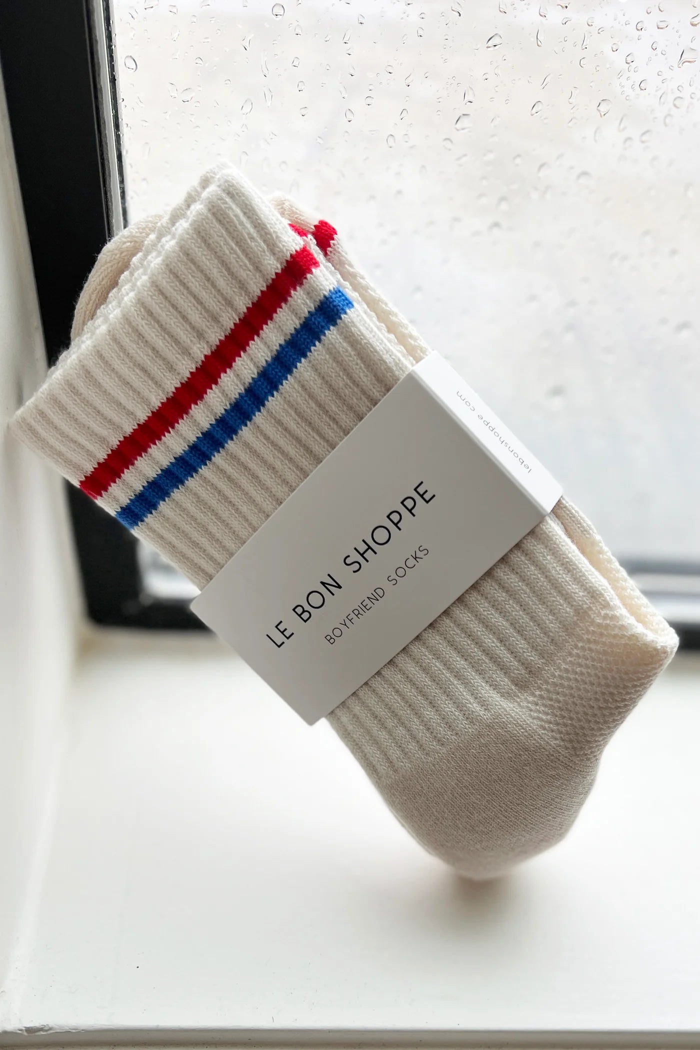 Le Bon Shoppe Boyfriend Socks in milk packaged with Le Bon Shoppe Boyfriend Socks band, propped against a rain-streaked window showing red and blue double 