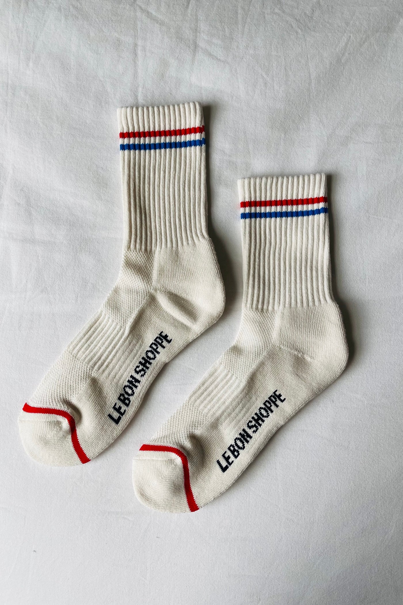 Pair of Le Bon Shoppe Boyfriend Socks in milk laid flat on white linen showing full longer-than-crew length, red and blue double stripe, Le Bon Shoppe sole 