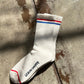 Single Le Bon Shoppe Boyfriend Sock in milk laid flat on concrete in dappled sunlight, showing red and blue double stripe, ribbed cotton texture and Le Bon Shoppe sole branding