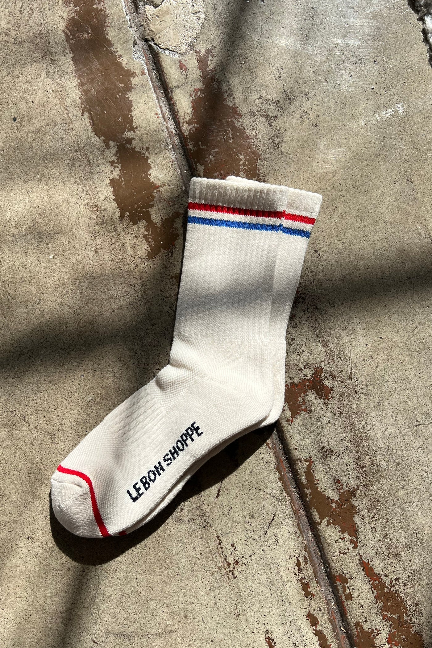 Single Le Bon Shoppe Boyfriend Sock in milk laid flat on concrete in dappled sunlight, showing red and blue double stripe, ribbed cotton texture and Le Bon Shoppe sole branding
