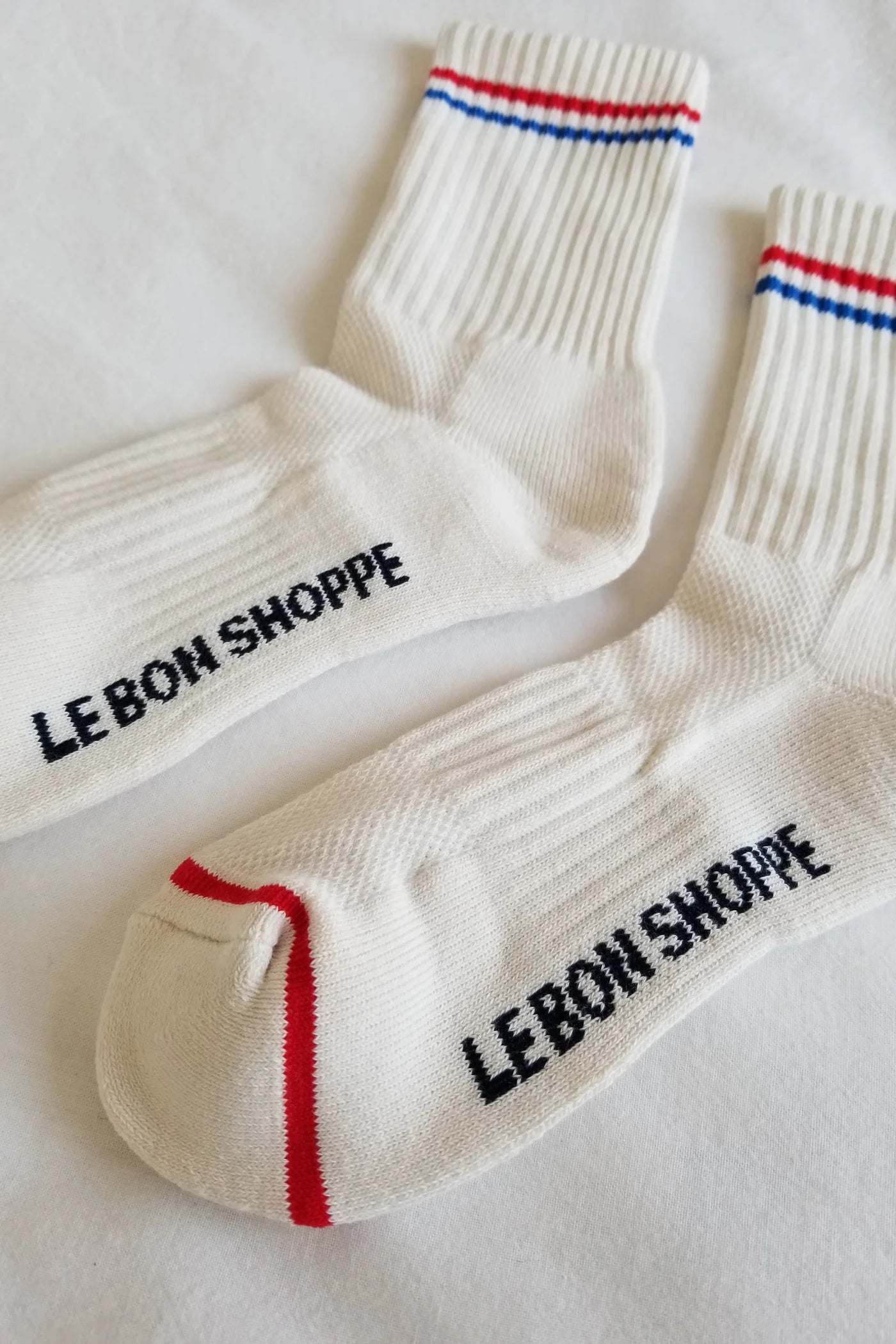 Close-up of Le Bon Shoppe Boyfriend Socks in milk showing sole branding, red contrast toe detail and red and blue double stripe on white ribbed cotton, laid on white linen