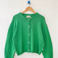 Le Bon Shoppe Cotton Leila Cardigan in spring green on a wooden hanger against a white wall, showing full relaxed silhouette, shell buttons and ribbed cotton knit detail