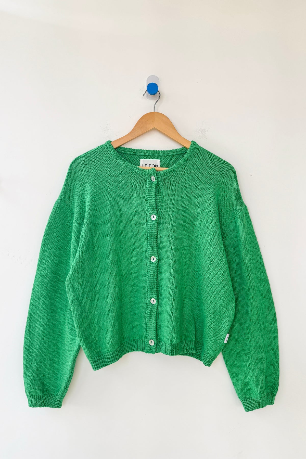 Le Bon Shoppe Cotton Leila Cardigan in spring green on a wooden hanger against a white wall, showing full relaxed silhouette, shell buttons and ribbed cotton knit detail