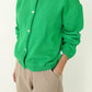 Three-quarter view of Le Bon Shoppe Cotton Leila Cardigan in spring green partially buttoned, showing shell buttons and relaxed cotton knit, styled with cream trousers
