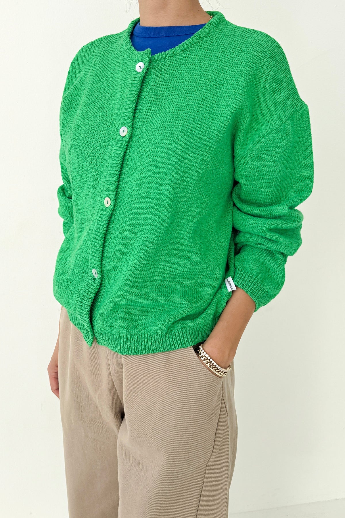 Three-quarter view of Le Bon Shoppe Cotton Leila Cardigan in spring green partially buttoned, showing shell buttons and relaxed cotton knit, styled with cream trousers