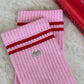 Close-up of Le Bon Shoppe Embroidered Boyfriend Socks in amour pink showing embroidered silver heart detail below red double stripe on pink ribbed cotton, laid on a marble surface
