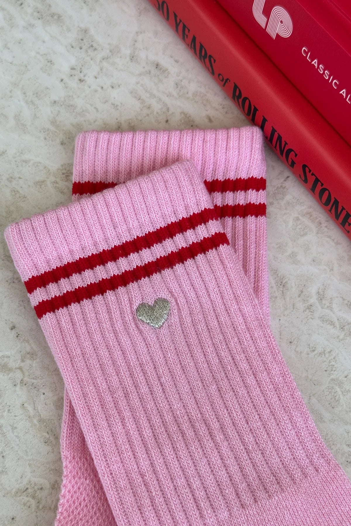 Close-up of Le Bon Shoppe Embroidered Boyfriend Socks in amour pink showing embroidered silver heart detail below red double stripe on pink ribbed cotton, laid on a marble surface
