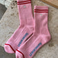 Pair of Le Bon Shoppe Embroidered Boyfriend Socks in amour pink laid flat on a travertine surface showing embroidered silver heart, red double stripe, sole branding and red contrast toe