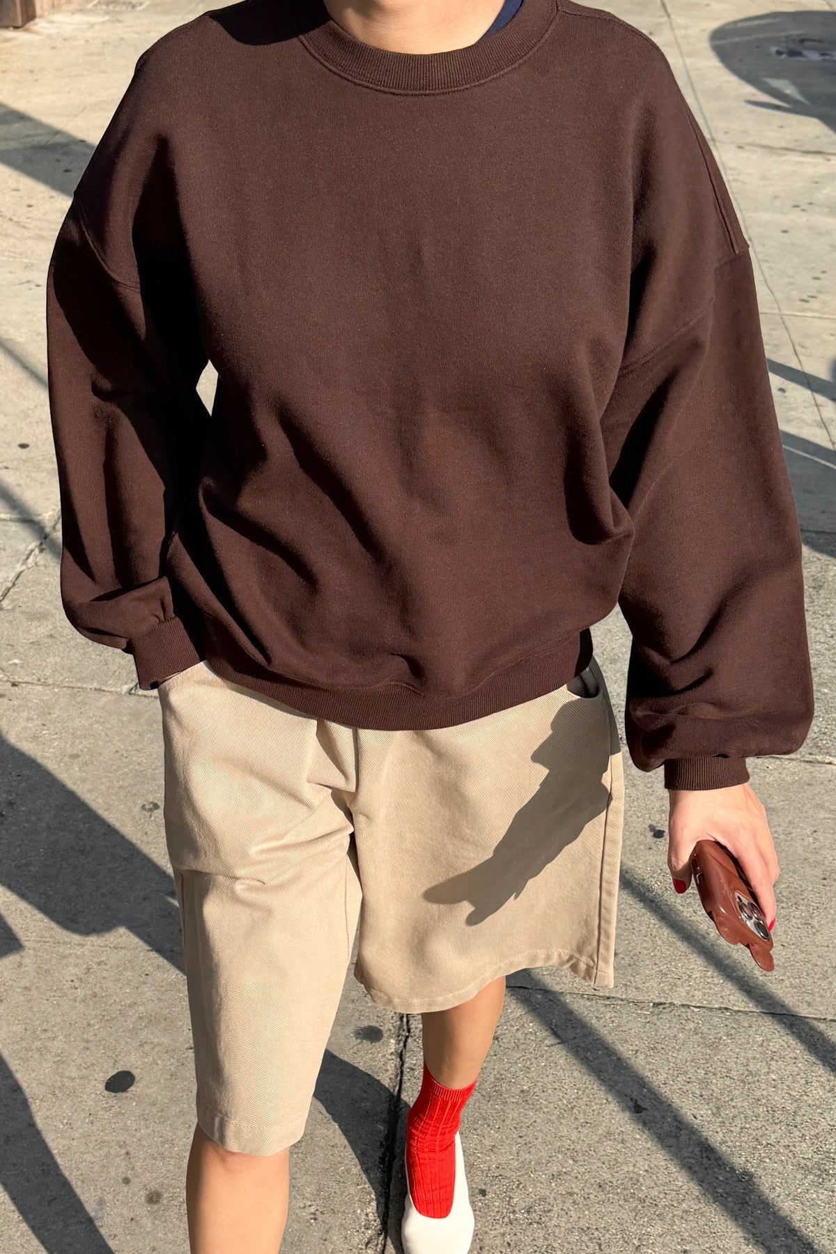 Le Bon Shoppe French Terry Core Sweatshirt in cocoa bean worn with taupe shorts, red ribbed socks and white shoes, shot on a sunny street