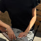 Le Bon Shoppe Go-To Tee in true black worn seated outdoors with black and white gingham trousers and a gold chain bracelet, showing relaxed fit in natural light