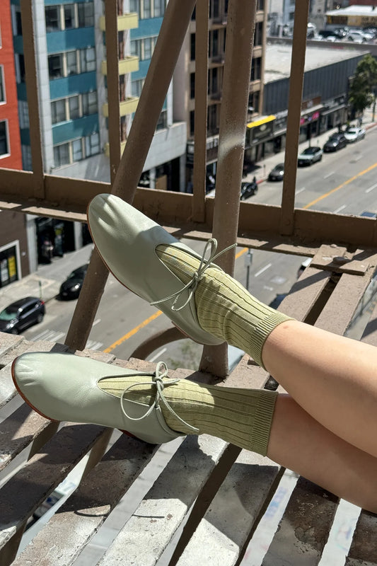 Le Bon Shoppe Her Socks in avocado cotton blend worn with sage green leather ballet flats, legs resting on a fire escape railing with a city street in the background