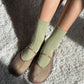 Le Bon Shoppe Her Socks in avocado green paired with brown Mary Jane shoes, standing on a cream shag rug with warm natural light and shadow