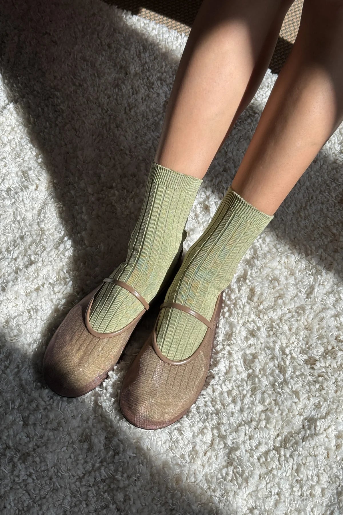 Le Bon Shoppe Her Socks in avocado green paired with brown Mary Jane shoes, standing on a cream shag rug with warm natural light and shadow