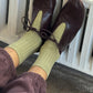 Le Bon Shoppe Her Socks in avocado green styled with dark burgundy leather oxford shoes and mauve trousers, resting on a white cast iron radiator