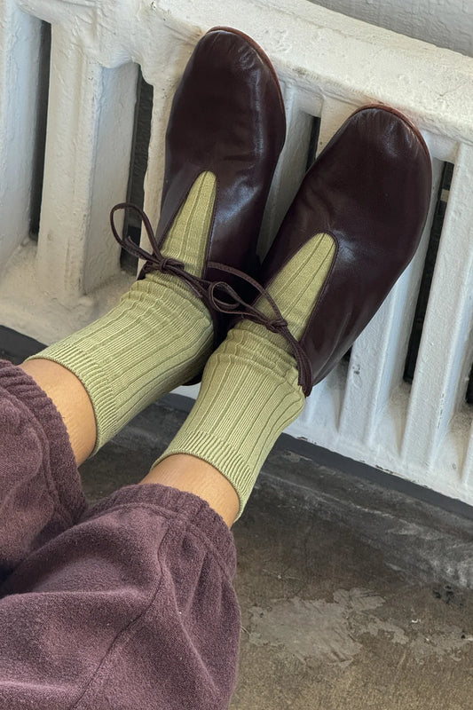 Le Bon Shoppe Her Socks in avocado green styled with dark burgundy leather oxford shoes and mauve trousers, resting on a white cast iron radiator