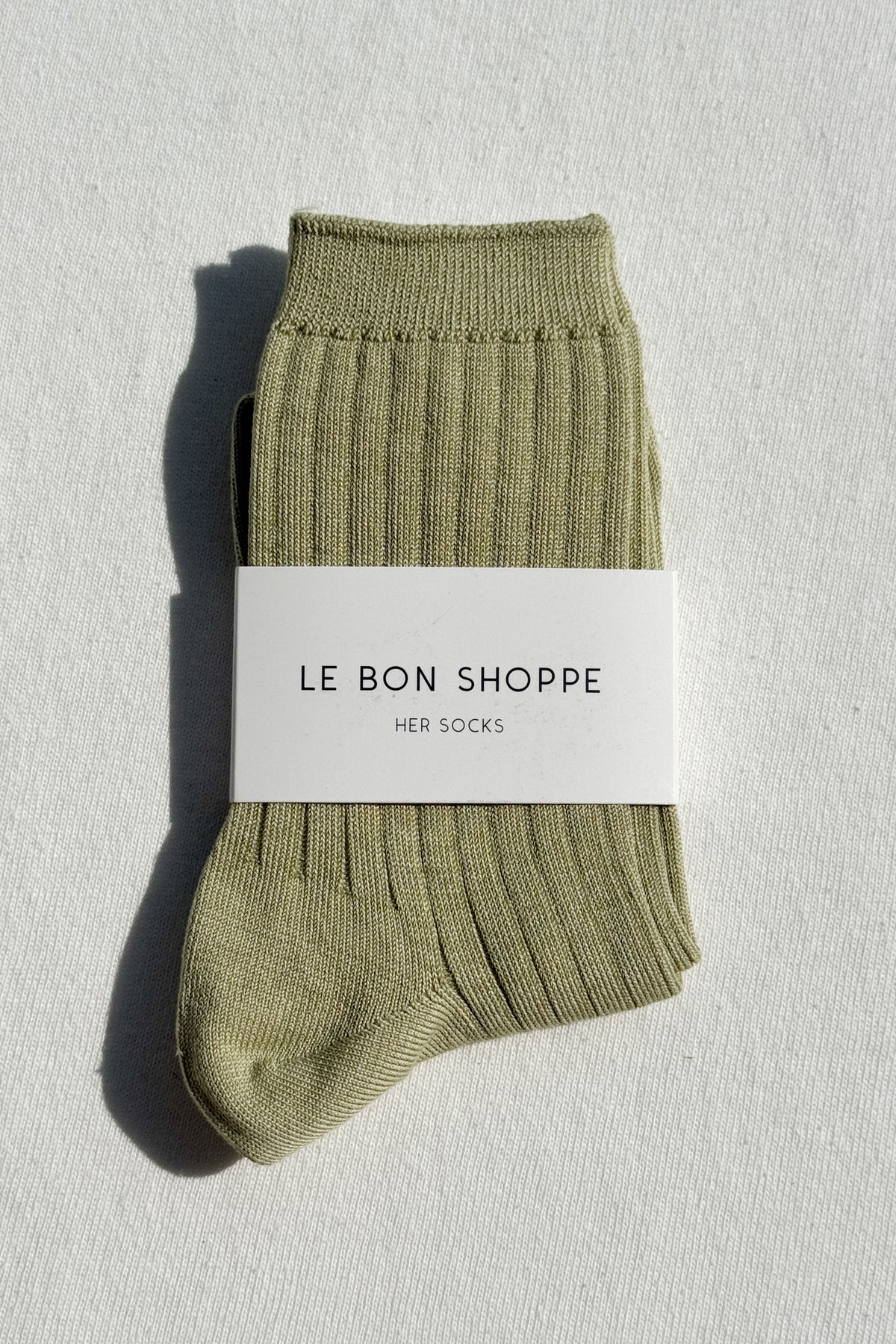 Le Bon Shoppe Her Socks in avocado green, folded and banded with branded packaging label, displayed on a white linen surface