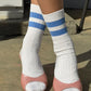 Le Bon Shoppe Her Varsity Socks in periwinkle worn with pink open-toe leather heels, showing white ribbed cotton-blend sock with periwinkle double stripe detail in natural sunlight