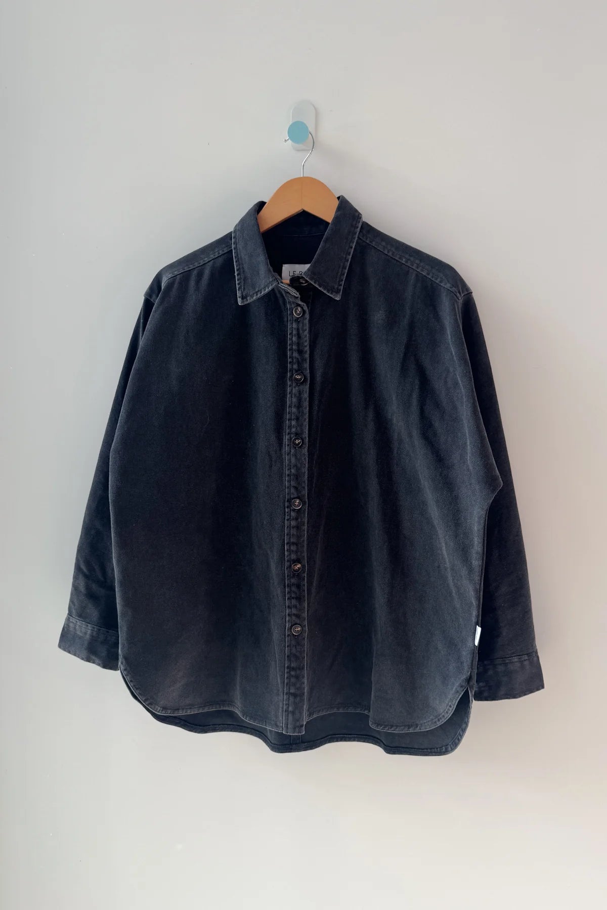 Le Bon Shoppe Theo Jacket in black denim on a wooden hanger against a grey wall, showing full oversized silhouette, button front and rounded hem