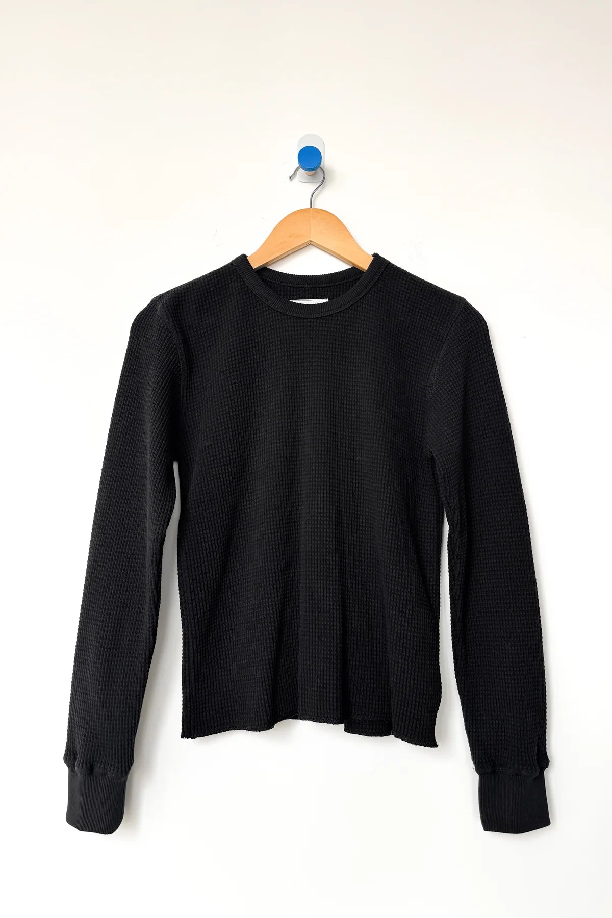 Le Bon Shoppe Waffle Fundamental Tee in true black on a wooden hanger against a white wall, showing full long-sleeve silhouette, crew neck and baby ribbed cuffs in heavyweight waffle cotton