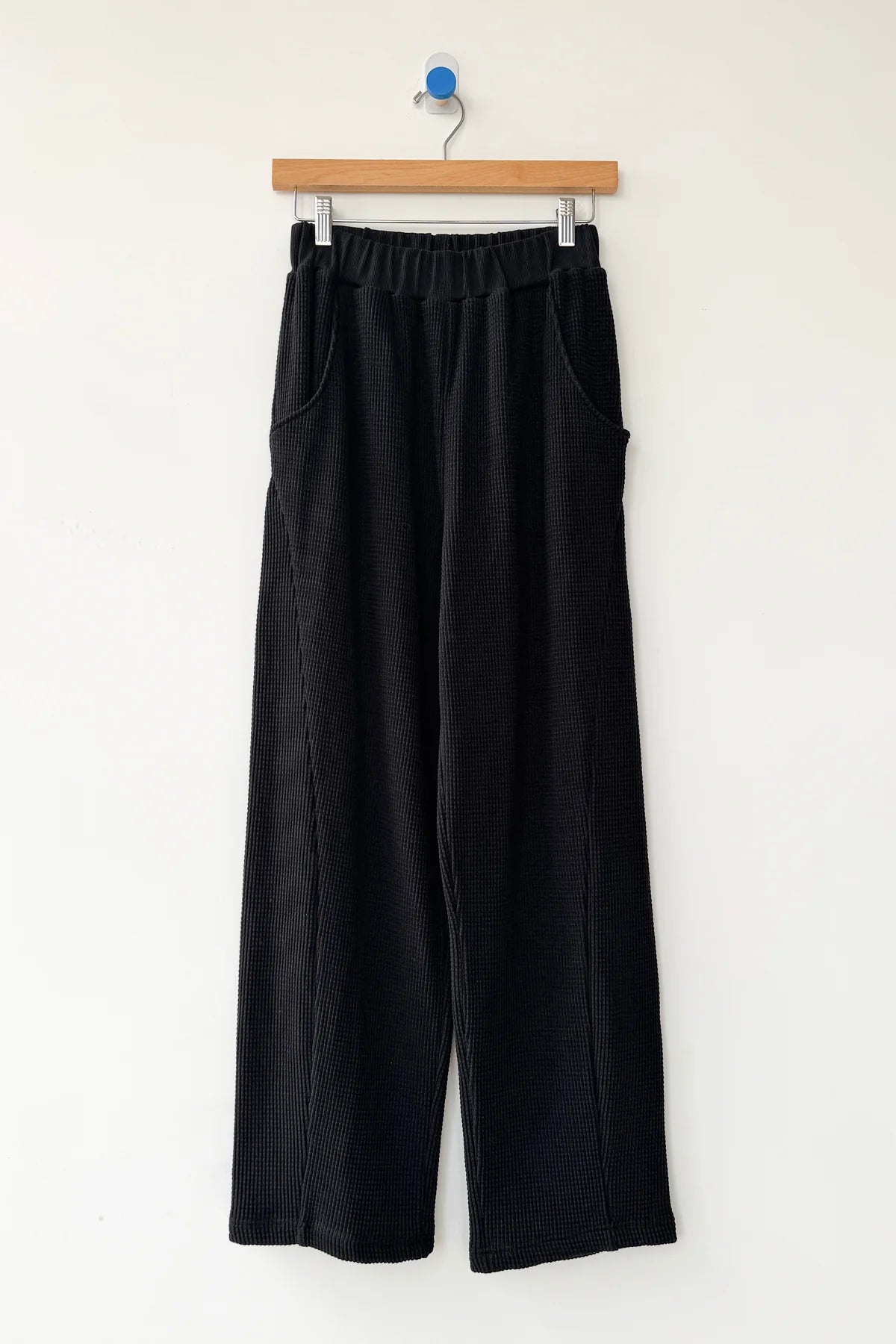Le Bon Shoppe Waffle Lo Pants in true black on a hanger against a white wall, showing full wide leg silhouette, elastic waistband, front pockets and signature arc seam in heavy waffle cotton